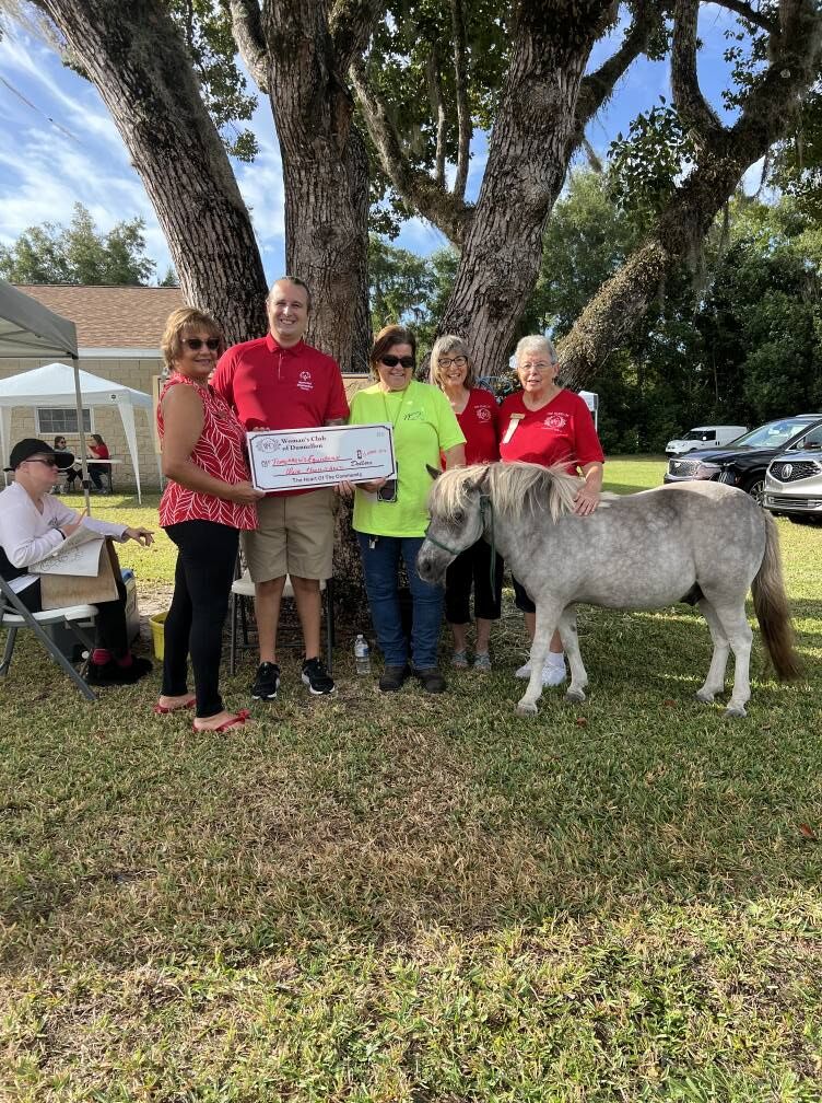 Members hand over a check on the lawn beside a miniature horse